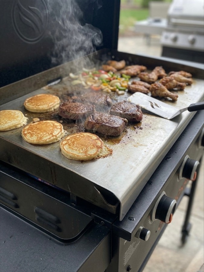 Steaks, wings, and pancakes cooking on a STELEBOARD used as a flat-top griddle over a gas grill